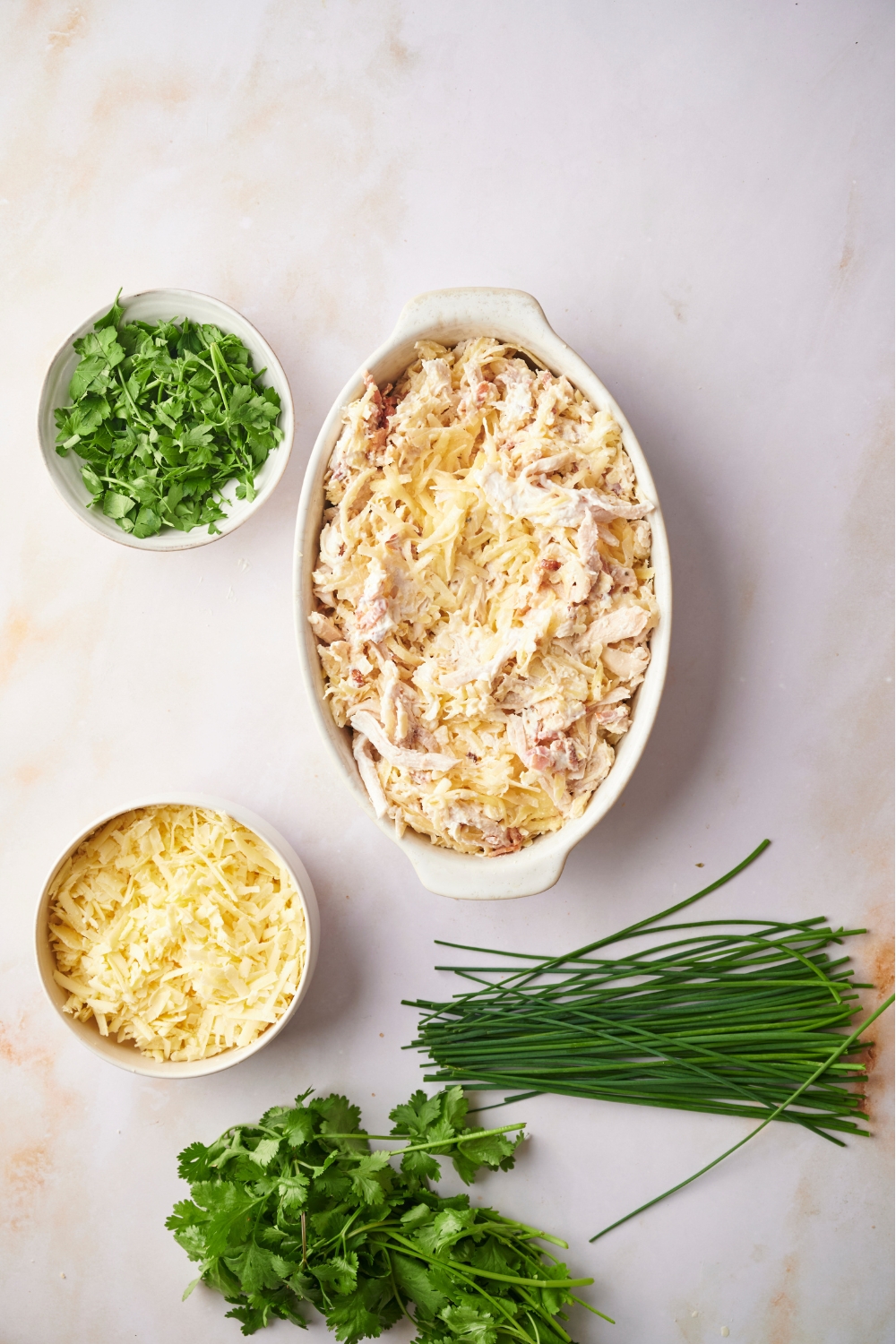 White casserole dish with unbaked chicken hashbrown casserole surrounded by ingredients including fresh herbs, chives, and shredded cheese.
