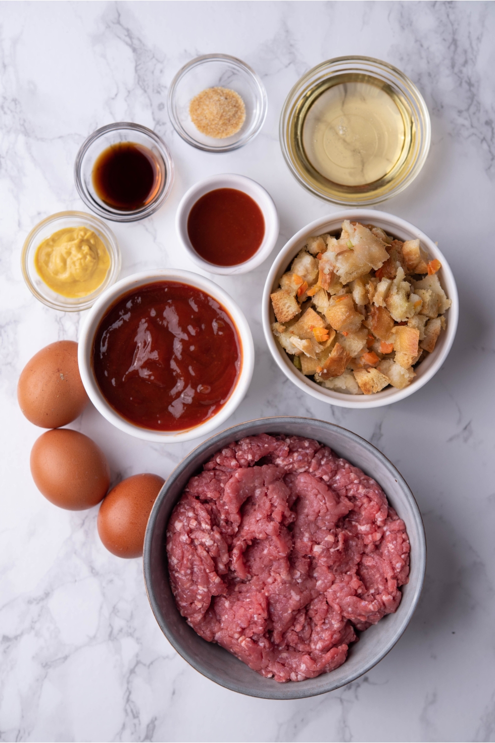 An assortment of ingredients including bowls of ground beef, ketchup, stuffing mix, oil, mustard, and spices.