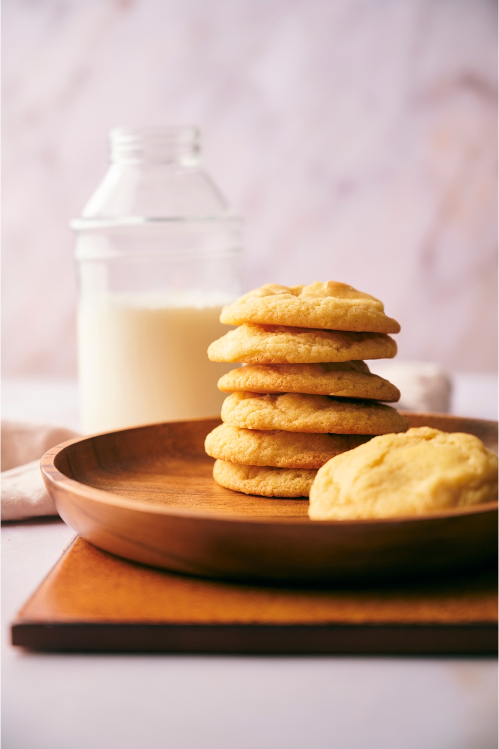 A stack of cookies with one fallen over on a wooden plate with a jar of milk in the background.