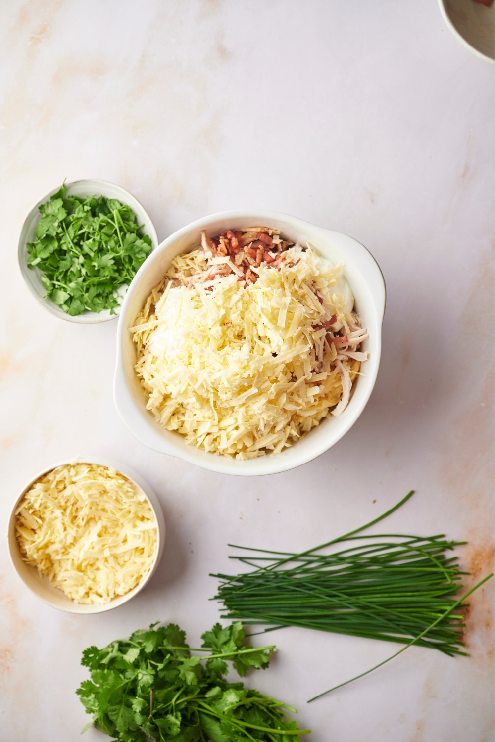 White mixing bowl filled with bacon, shredded cheese, chicken, uncooked hashbrowns, and cream cheese. Next to the bowl are fresh herbs and a bowl of shredded cheese.