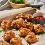 Baking sheet lined with parchment paper with sausage balls garnished with fresh herbs and a small bowl of ketchup on the baking sheet.