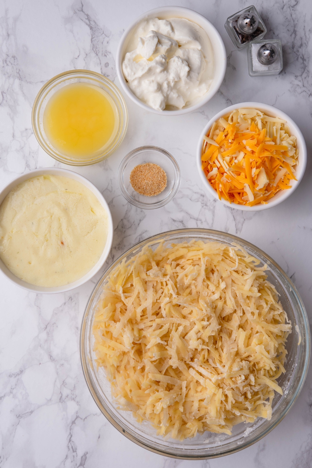 A countertop with a bowl of thawed shredded hash browns, sour cream, shredded cheese, cream of chicken soup, melted butter, pepper, and onion powder.