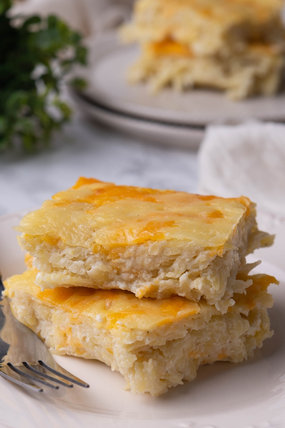 A stack of two squares of hash brown casserole on a plate with a fork next to it.