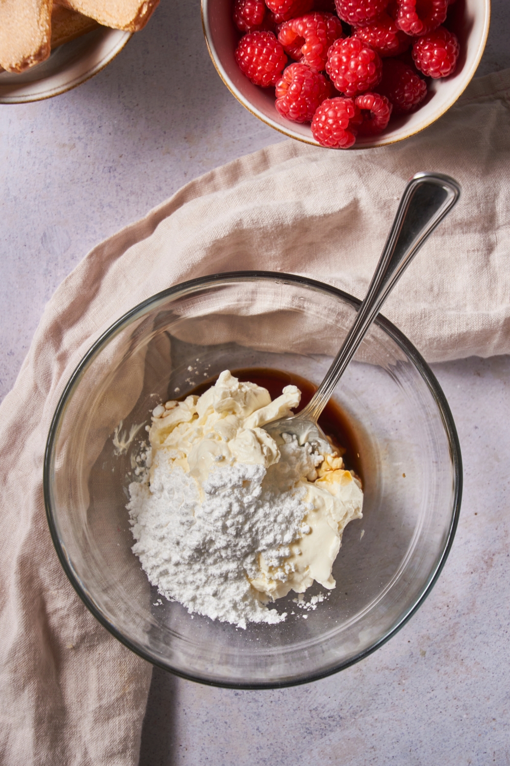 A clear mixing bowl with a spoon and the bowl is filled with powdered sugar, cream cheese, and vanilla extract, none of which has been mixed together.