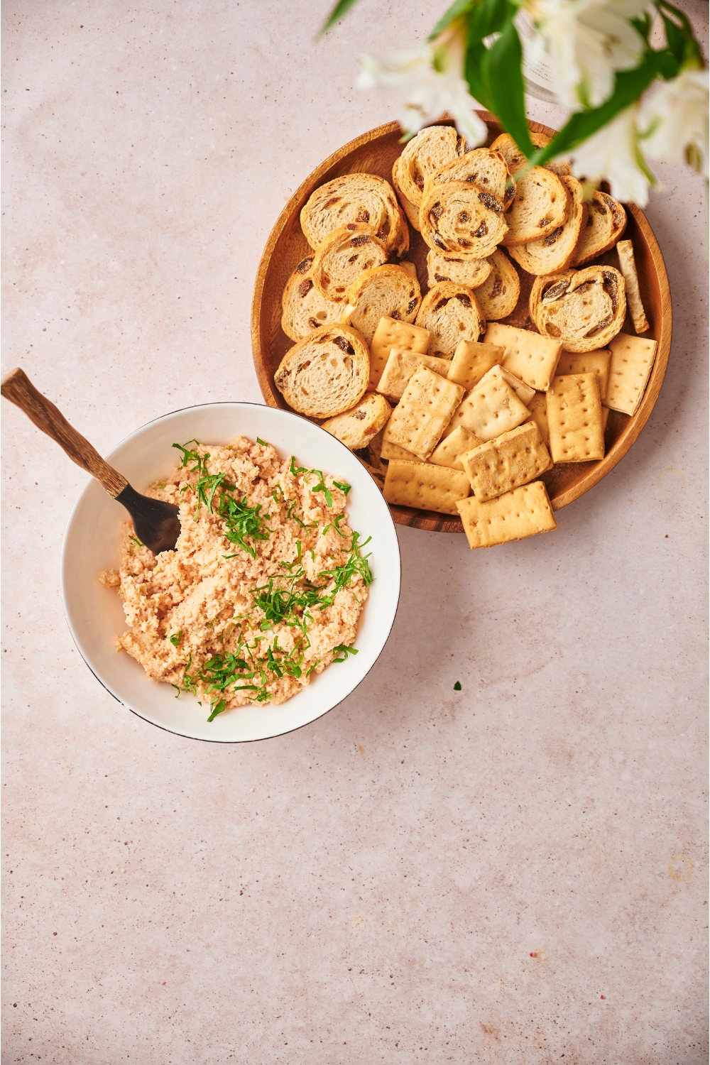 Deviled ham topped with fresh herbs in a white bowl with a spoon, next to a plate of crackers and bread slices.