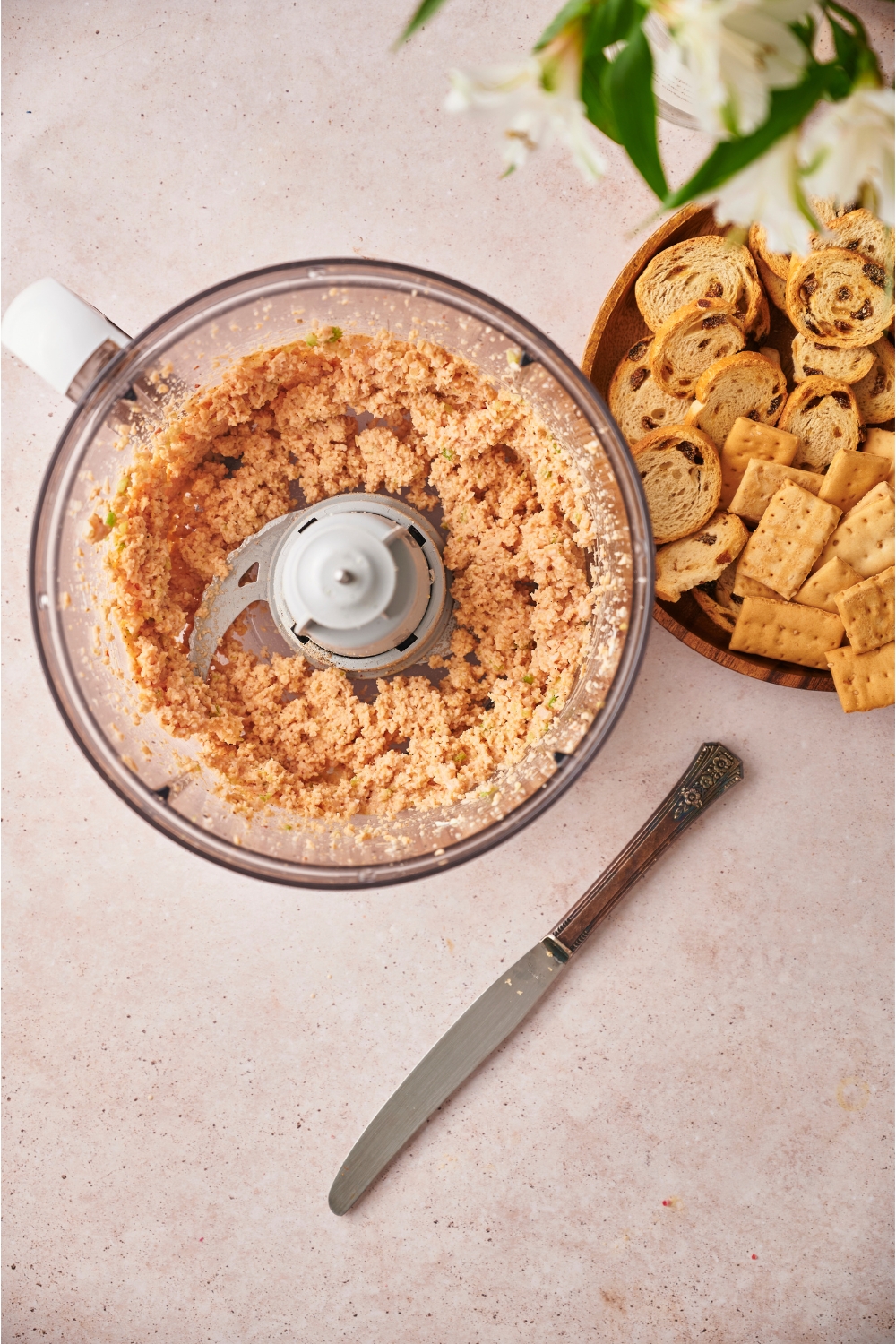 Food processor with processed ham mixture next to a knife and a plate of crackers and bread.