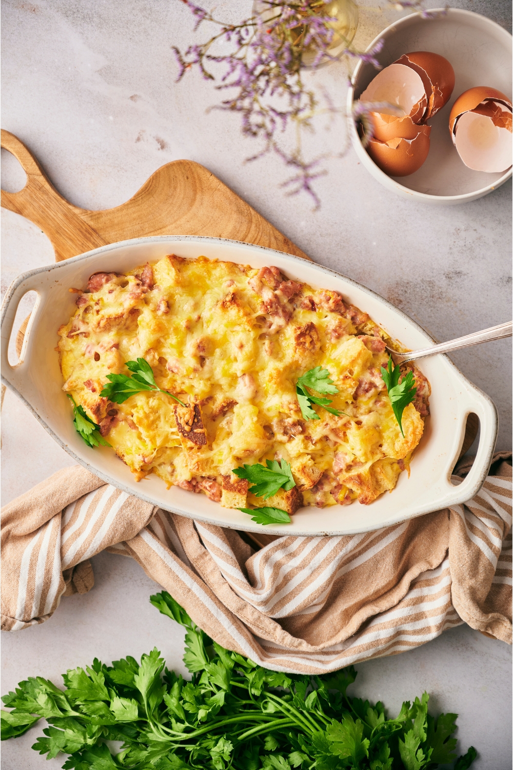 Christmas breakfast casserole in a white casserole dish with a serving spoon in the dish. The dish is on a wood board with a kitchen towel next to the dish.