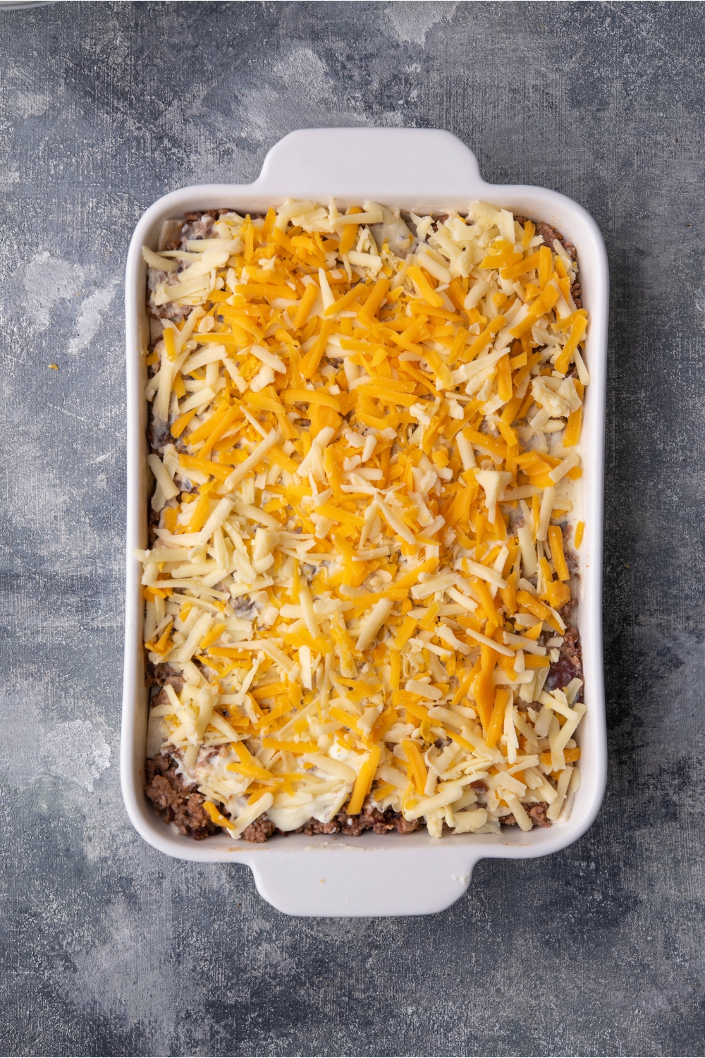 White baking dish with unbaked burrito casserole covered in shredded cheese.