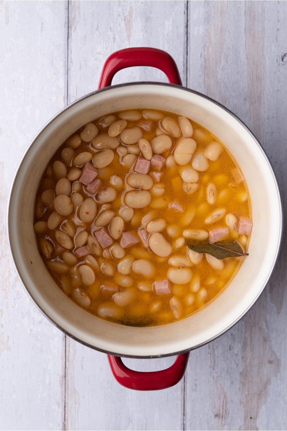 A red sauce pot filled with butter beans, diced ham and bay leaves in a seasoned broth.