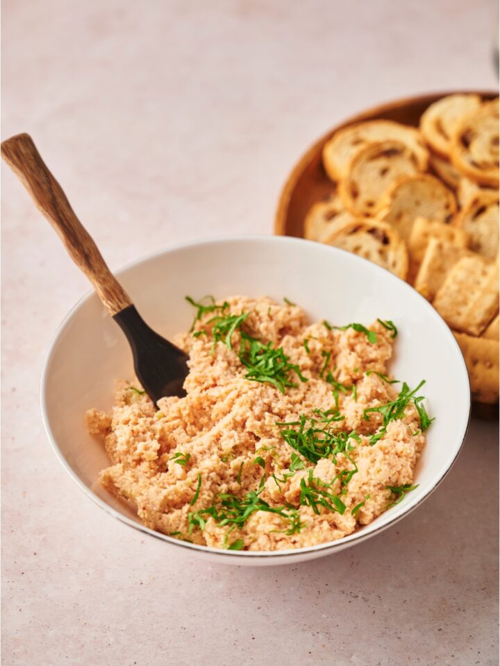 Deviled ham topped with fresh herbs in a white bowl with a spoon, next to a plate of crackers and bread slices.