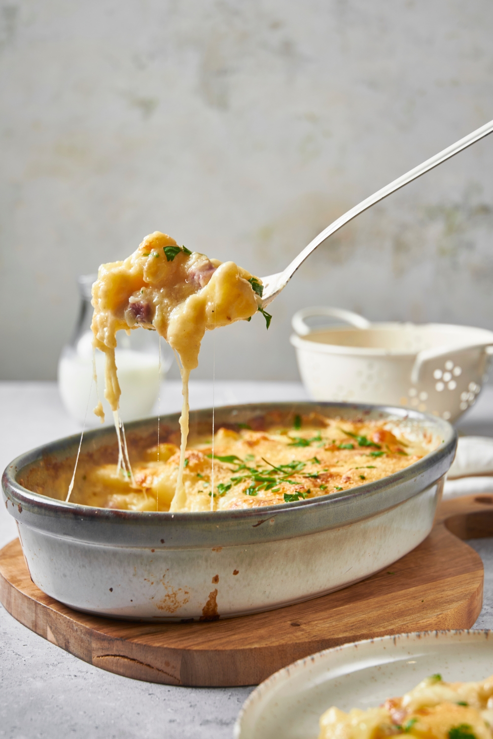 Casserole dish on a wooden board with a scoop of ham and potato casserole being scooped out of the dish by a large serving spoon.