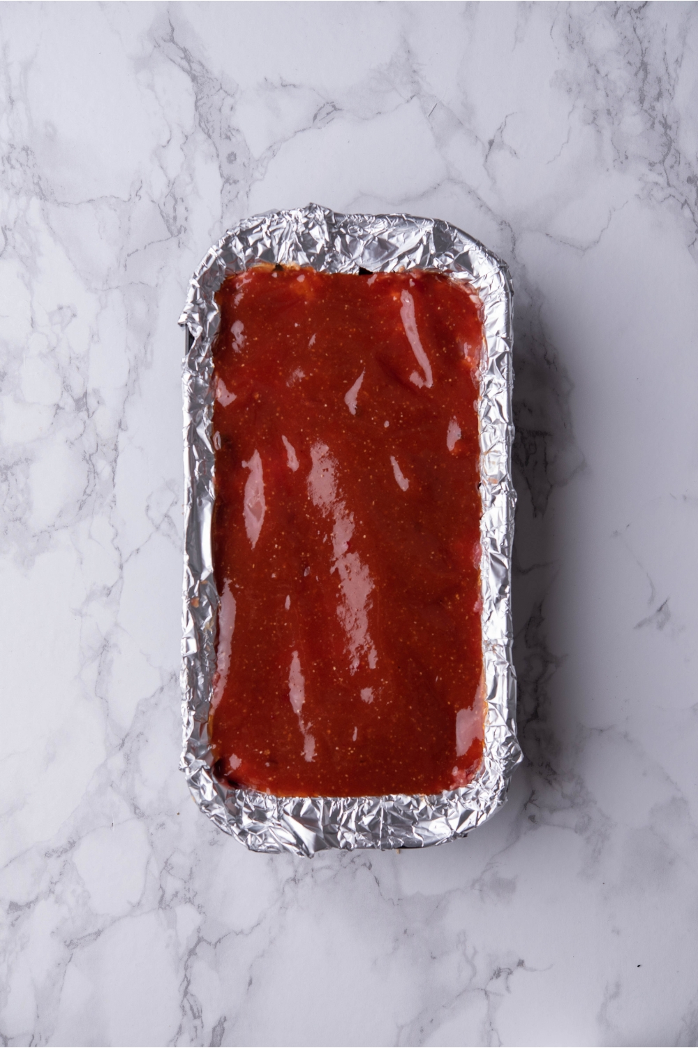 Loaf pan lined with aluminum foil filled with unbaked meatloaf covered in a ketchup glaze.