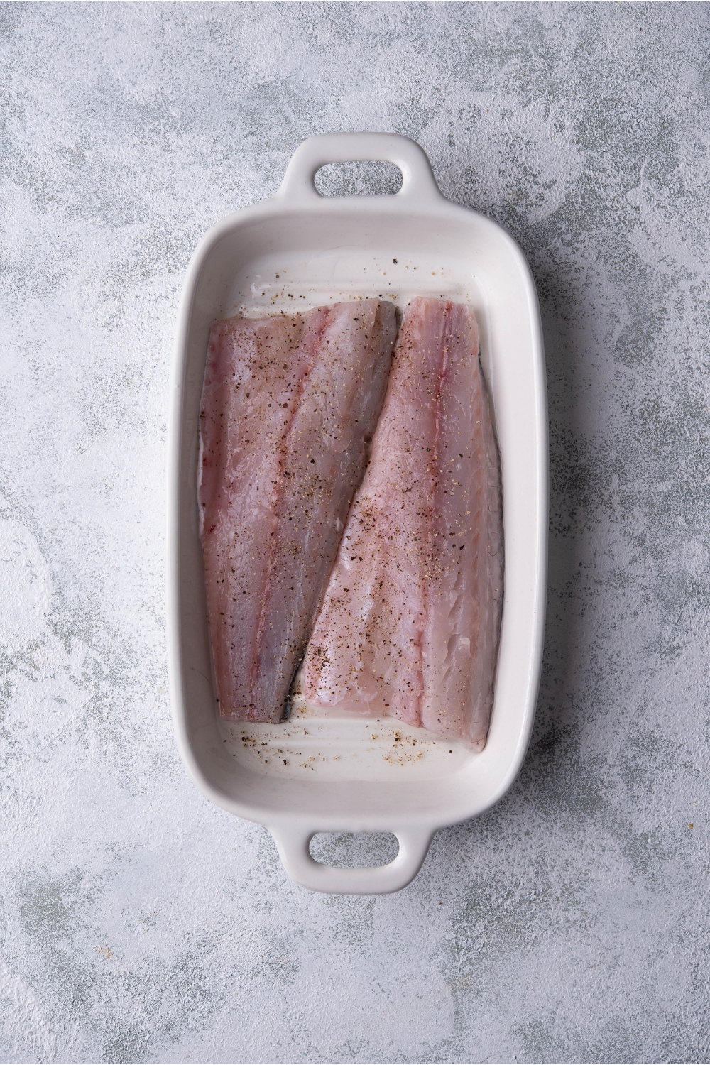 Two raw fish fillets seasoned with pepper in a white baking dish.