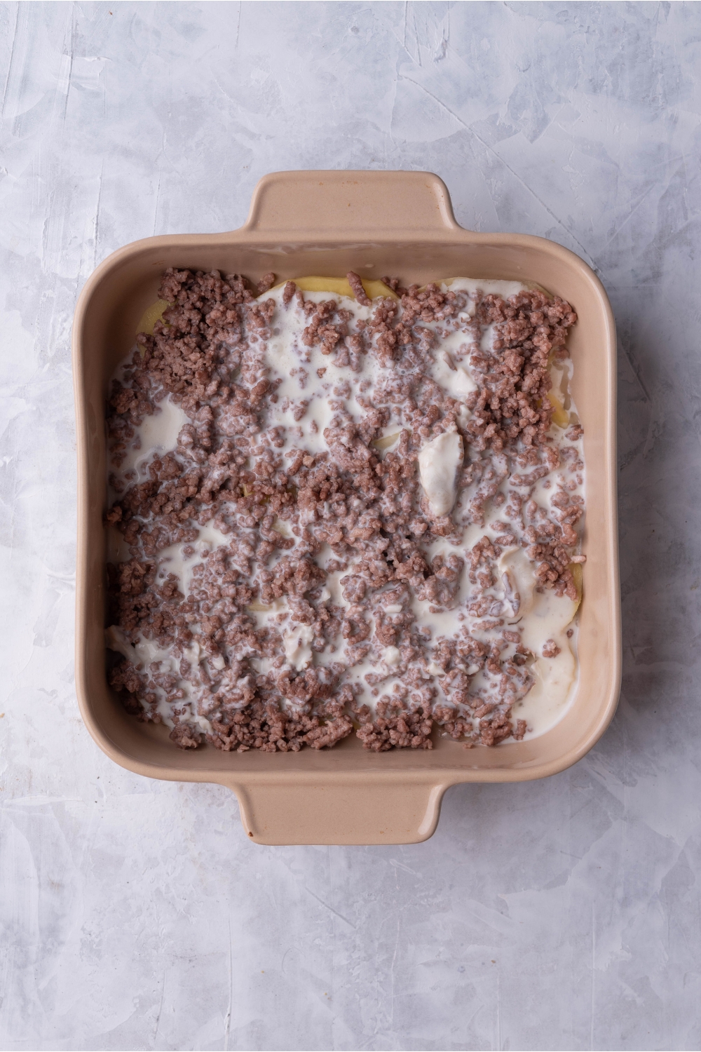 Brown baking dish with layers of cooked ground beef and a creamy sauce mixture.