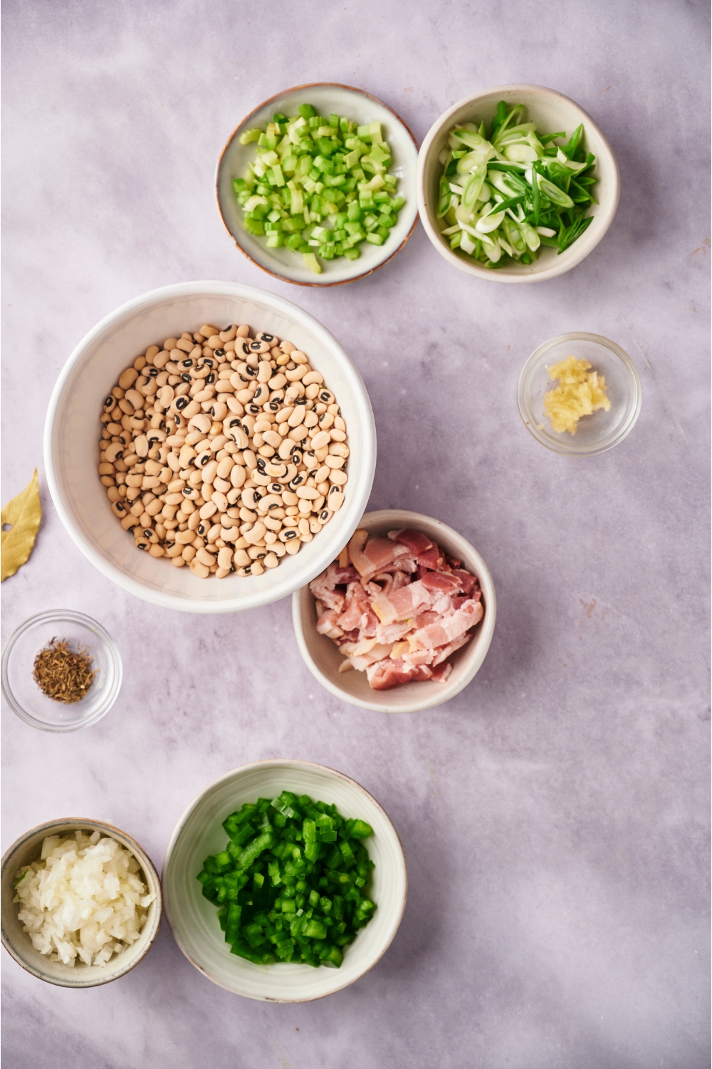 An assortment of ingredients including bowls of black eyed peas, bacon, green onion, peppers, onion, garlic, and herbs.