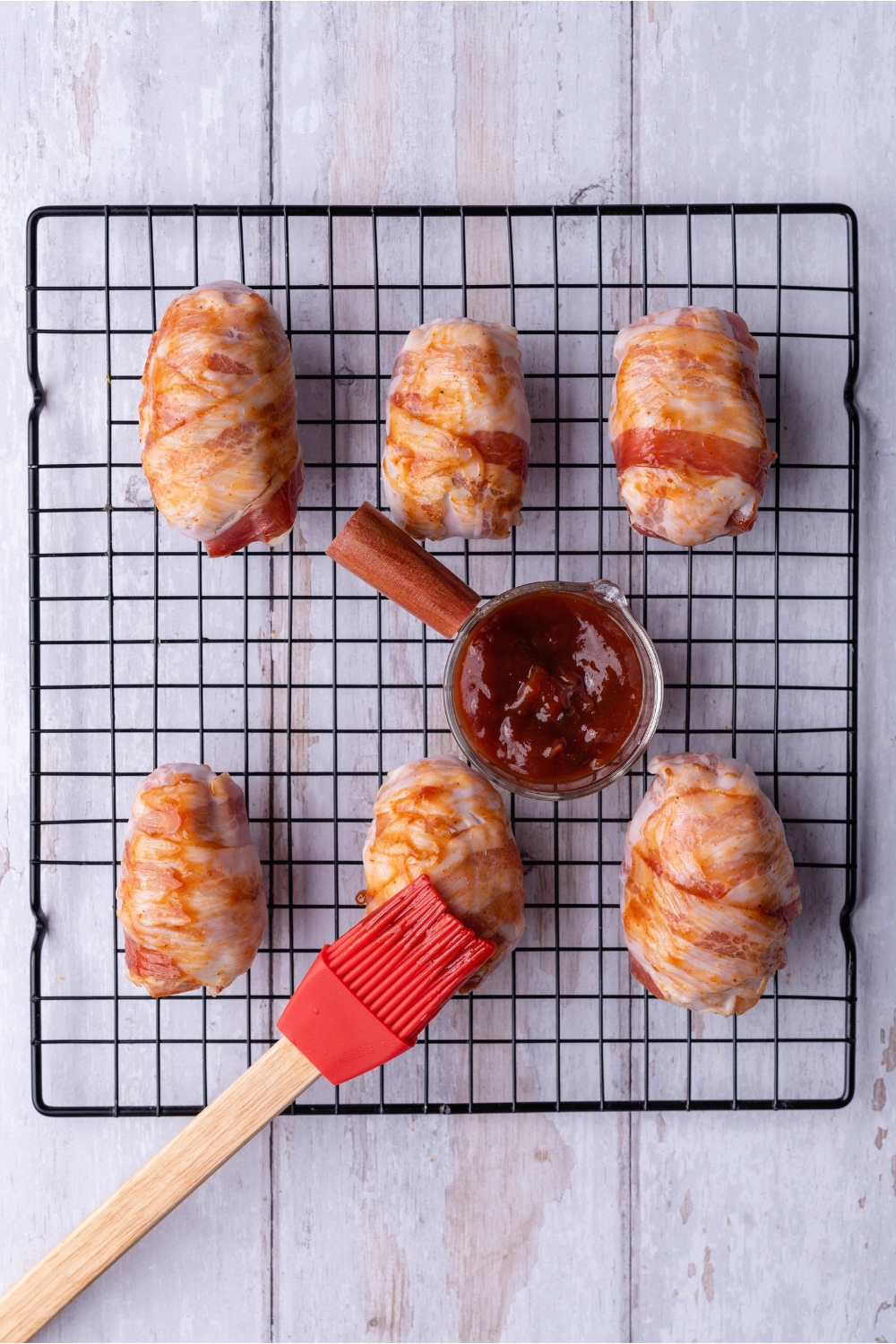 A wire rack with six bacon-wrapped armadillo eggs. A small bowl of BBQ sauce and a grill brush are on the rack.