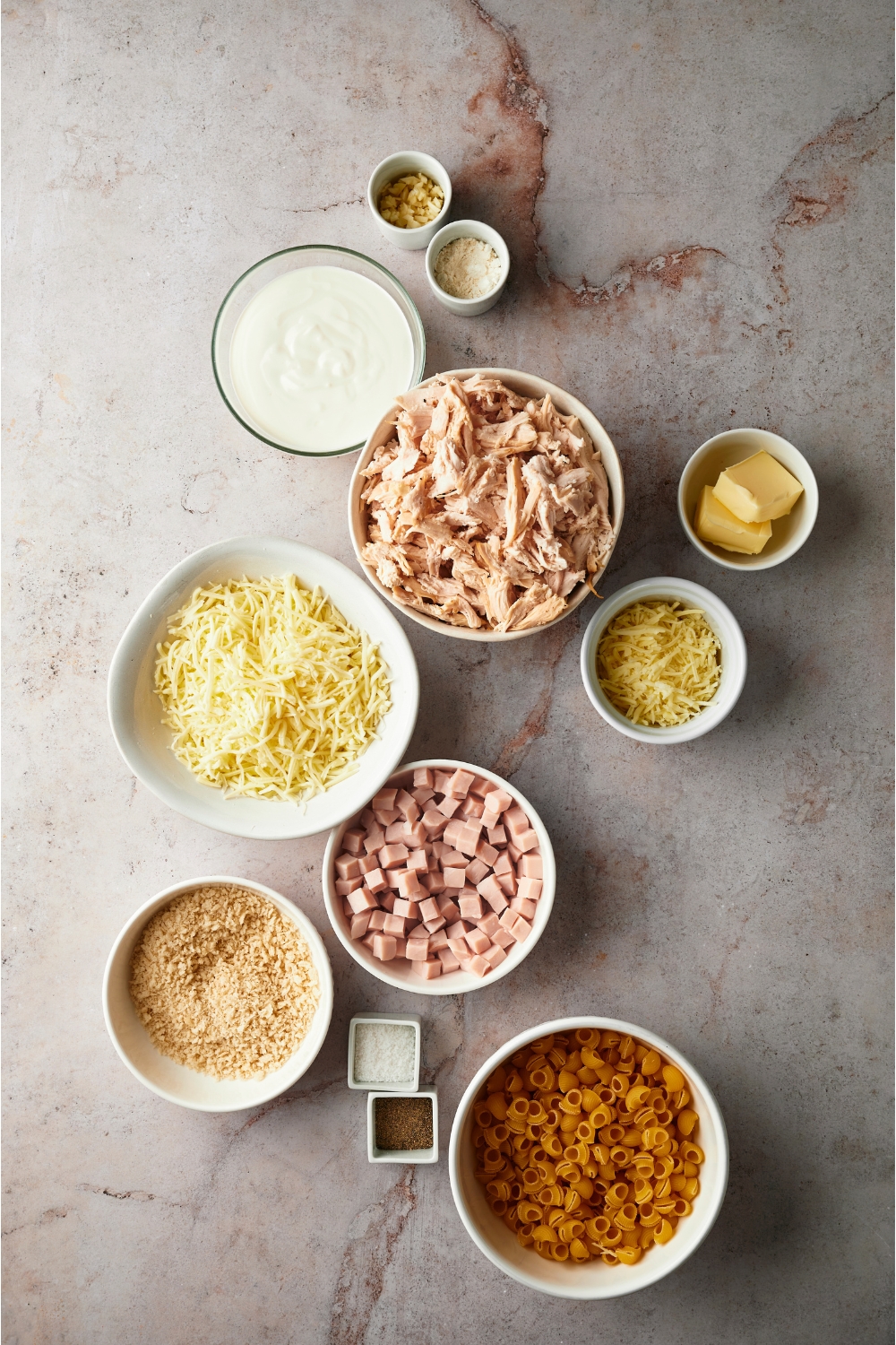 An assortment of ingredients for chicken cordon bleu casserole including bowls of shredded cheese, shredded chicken, uncooked pasta, bread crumbs, diced ham, butter and spices.