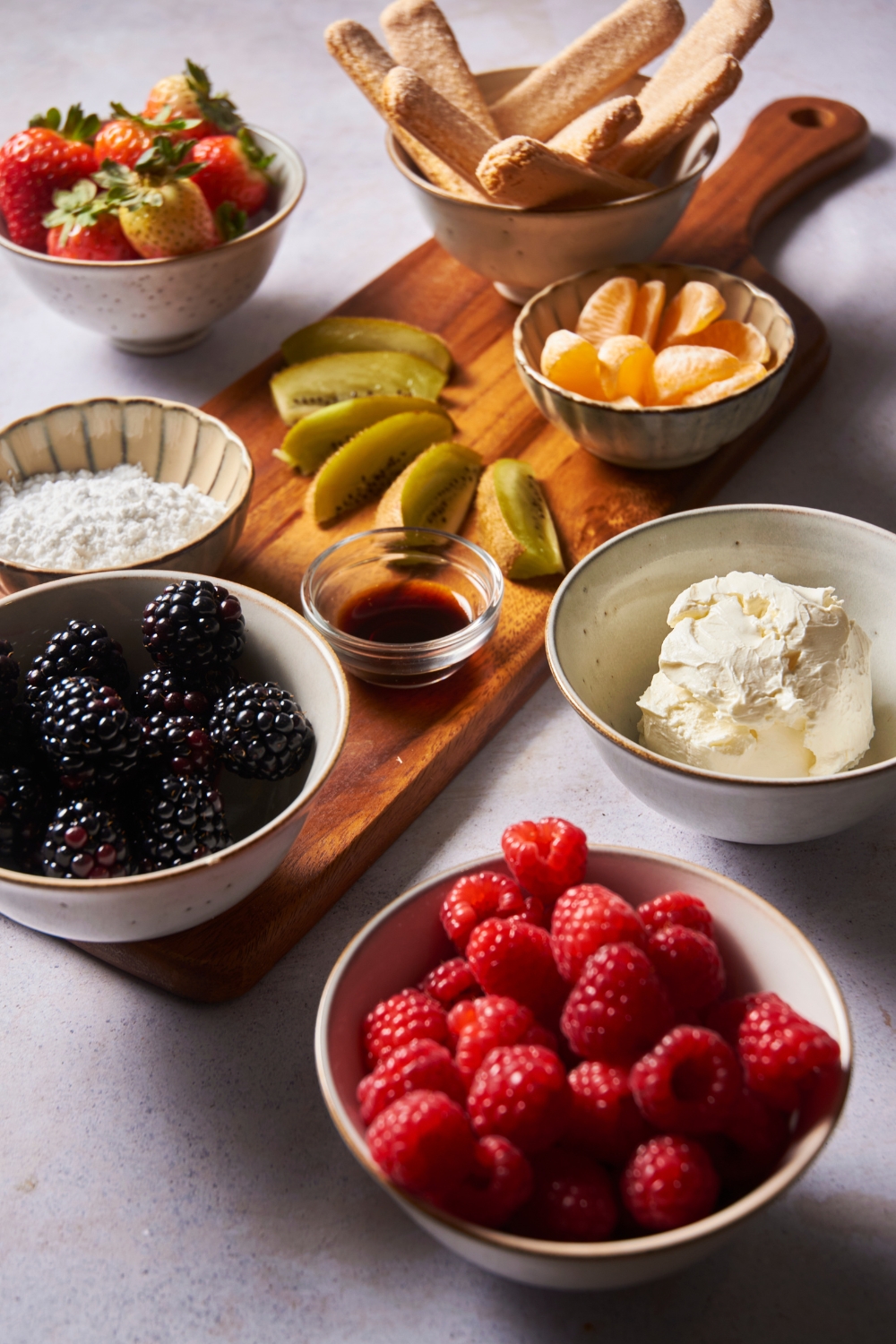 An assortment of ingredients including bowls of fresh raspberries, blackberries, cream cheese, tangerines, powdered sugar, and vanilla extract. Some of the ingredients are on top of a wooden board, others are on the counter.