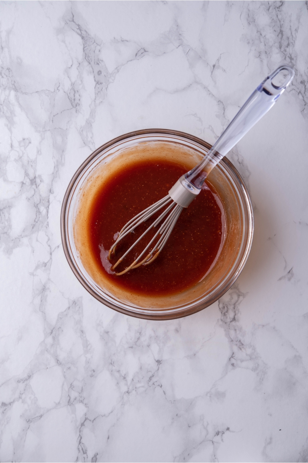 A clear bowl filled with a ketchup mixture that has been mixed with a whisk. The whisk is in the bowl.