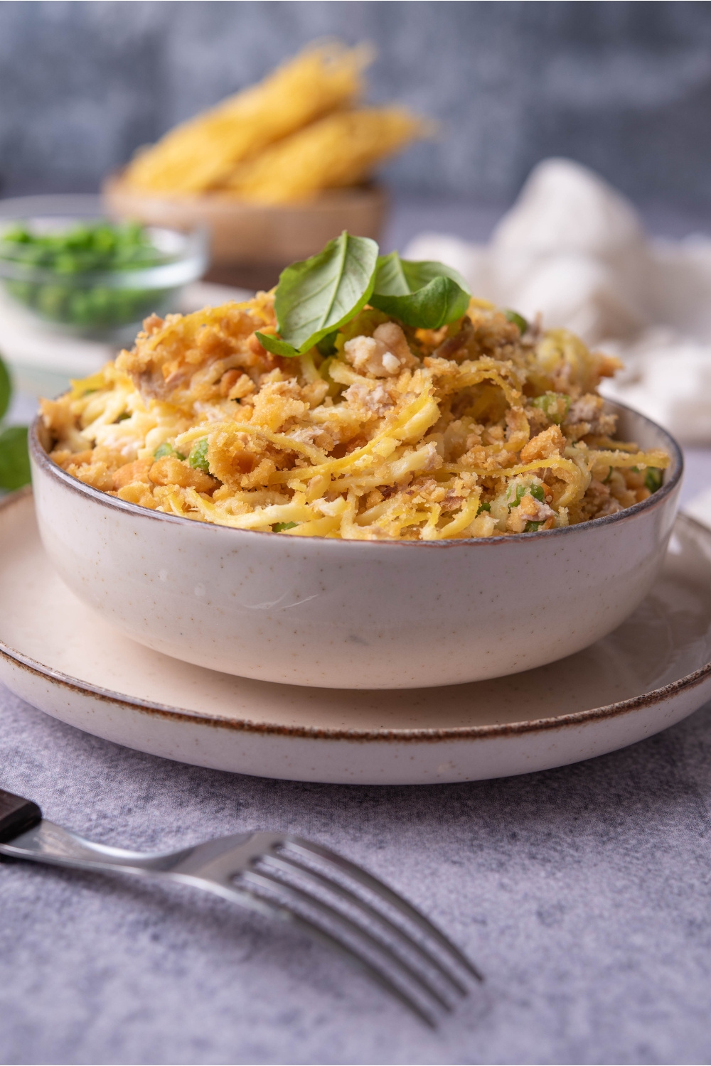 White bowl with a serving tuna noodle casserole on top of a white plate with a fork next to it.