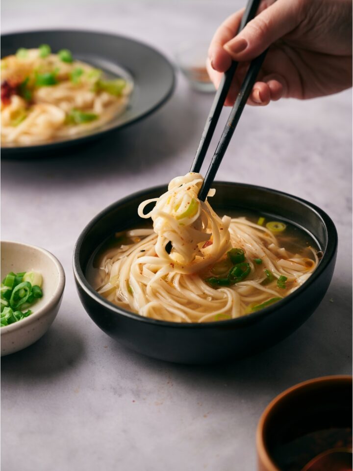 Hand holding chopsticks with a serving of noodles being taken from a black bowl of udon soup.