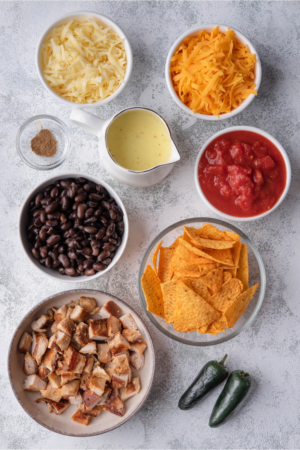 An assortment of ingredients including bowls of diced chicken, black beans, tortilla chips, crushed tomatoes, cheese, soup, and spices.