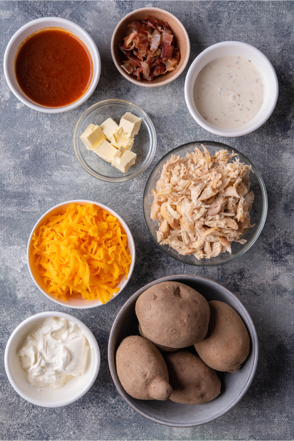 An assortment of ingredients for buffalo chicken casserole including bowls of potatoes, shredded cheese, shredded chicken, ranch dressing, buffalo sauce, and sour cream.