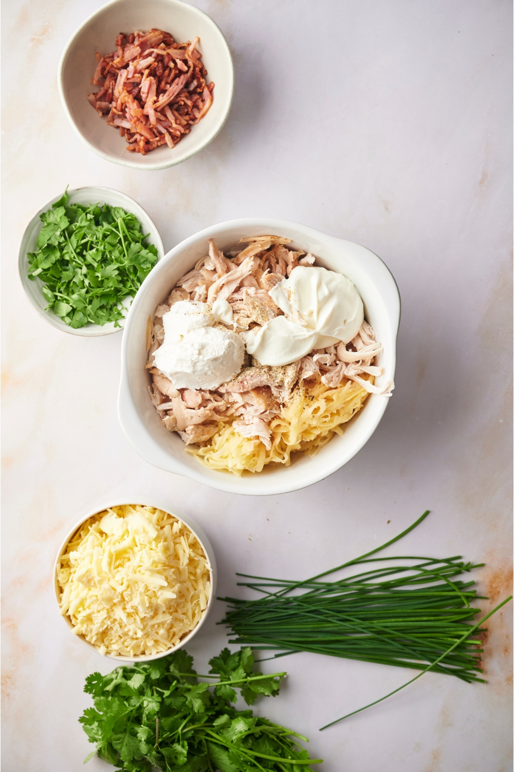 White mixing bowl filled with shredded chicken, cream cheese, spices, and uncooked hashbrowns, surrounded by bowls of other ingredients including bacon, fresh herbs, and shredded cheese.