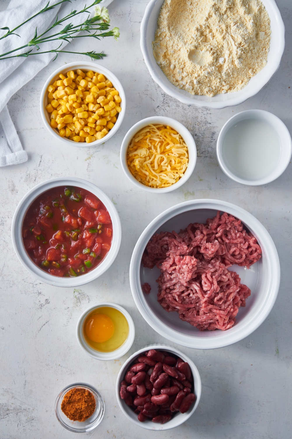 An assortment of ingredients including bowls of cornbread mix, raw ground beef, salsa, corn, shredded cheese, beans, an egg, and spices.