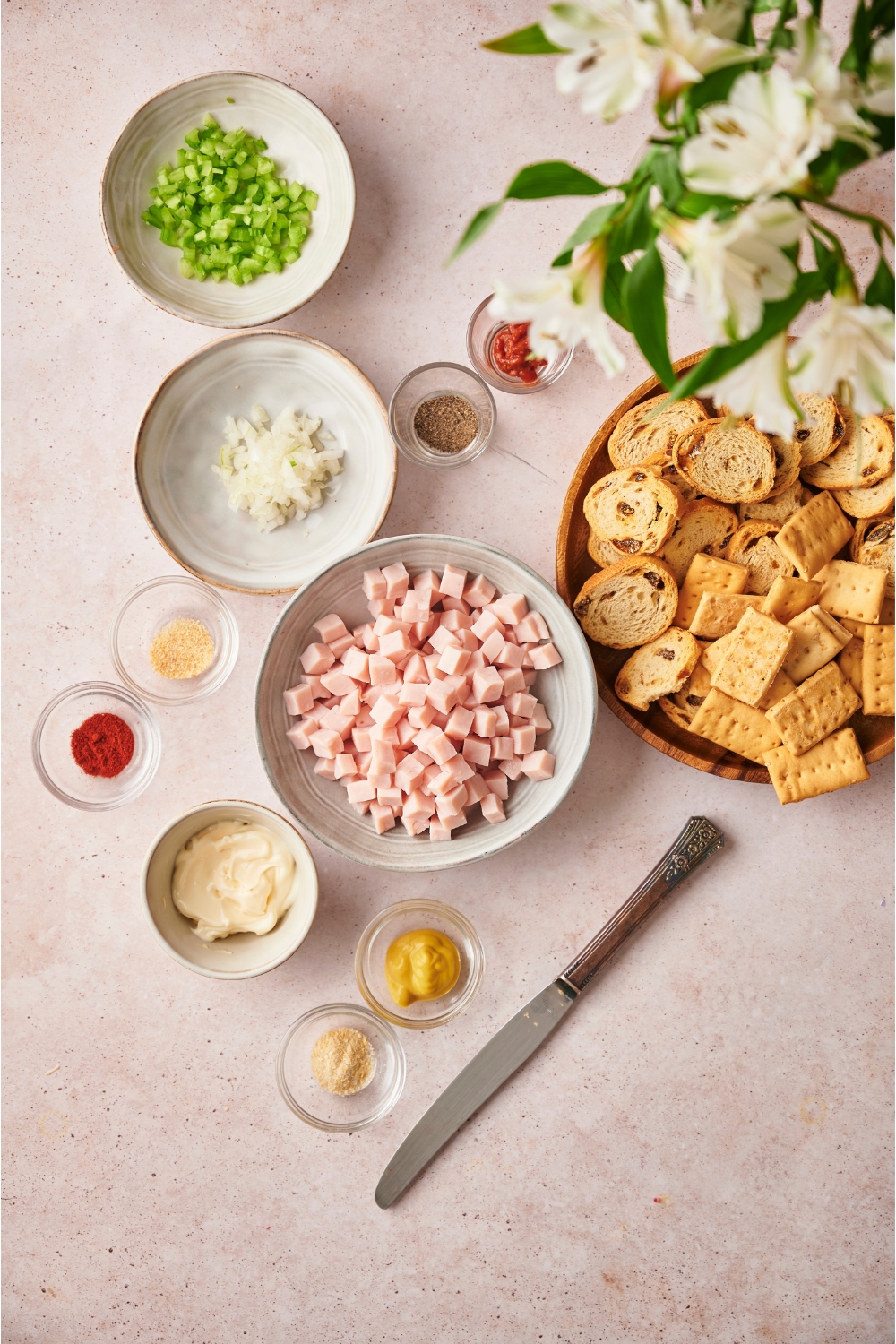 An assortment of ingredients including bowls of cubed ham, mayonnaise, diced celery, diced onion, mustard, spices, and a plate of crackers and bread.
