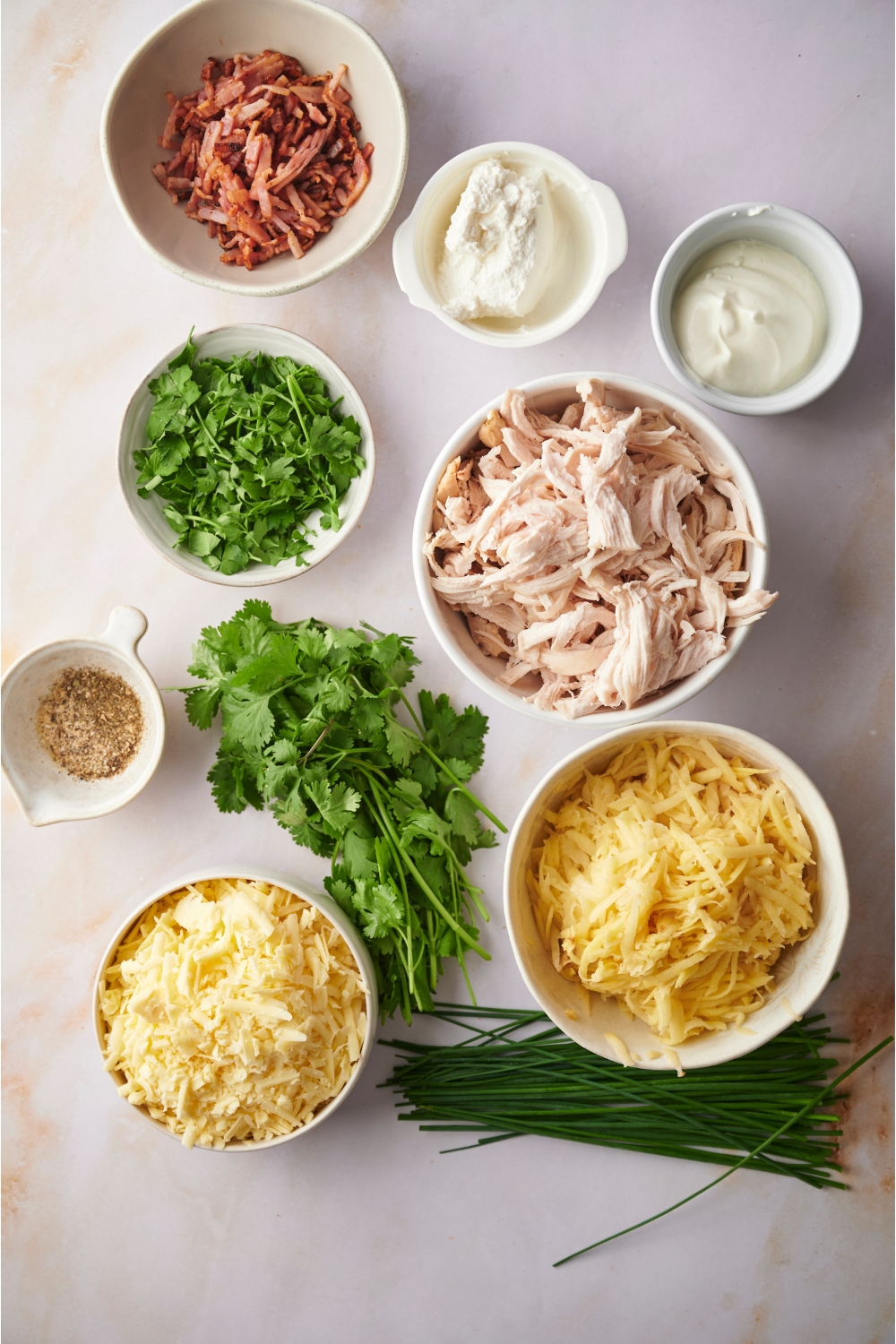 An assortment of ingredients including bowls of shredded chicken, uncooked hashbrowns, fresh herbs, shredded cheese, bacon, cream cheese, and sour cream.
