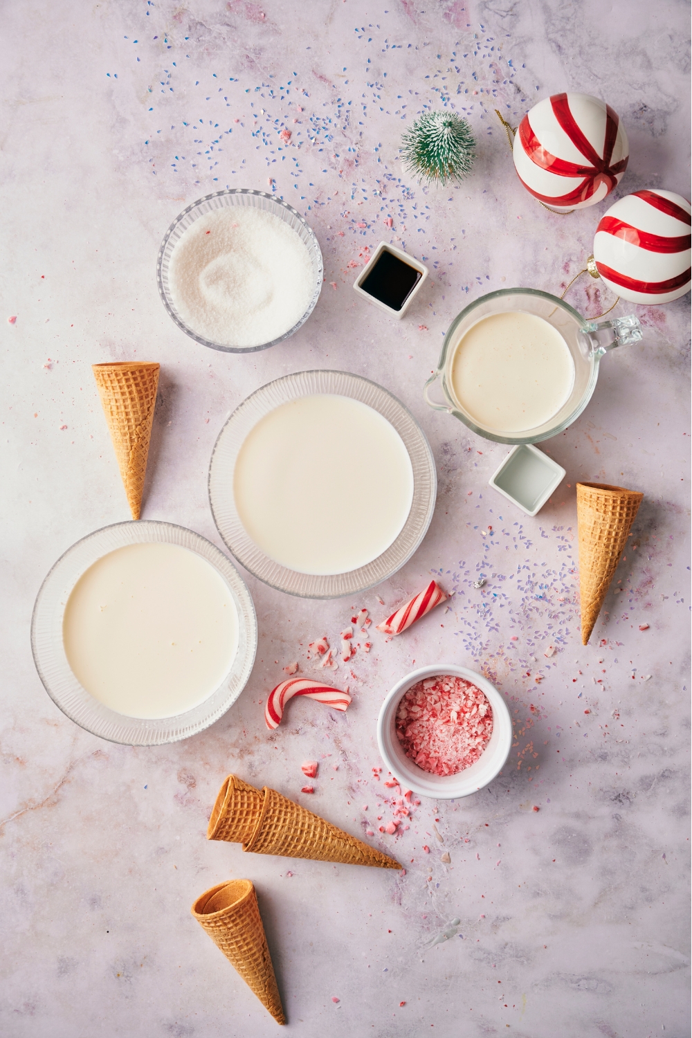 An assortment of ingredients including bowls of milk, cream, vanilla extract, sugar, ice cream cones, and crushed candy canes.