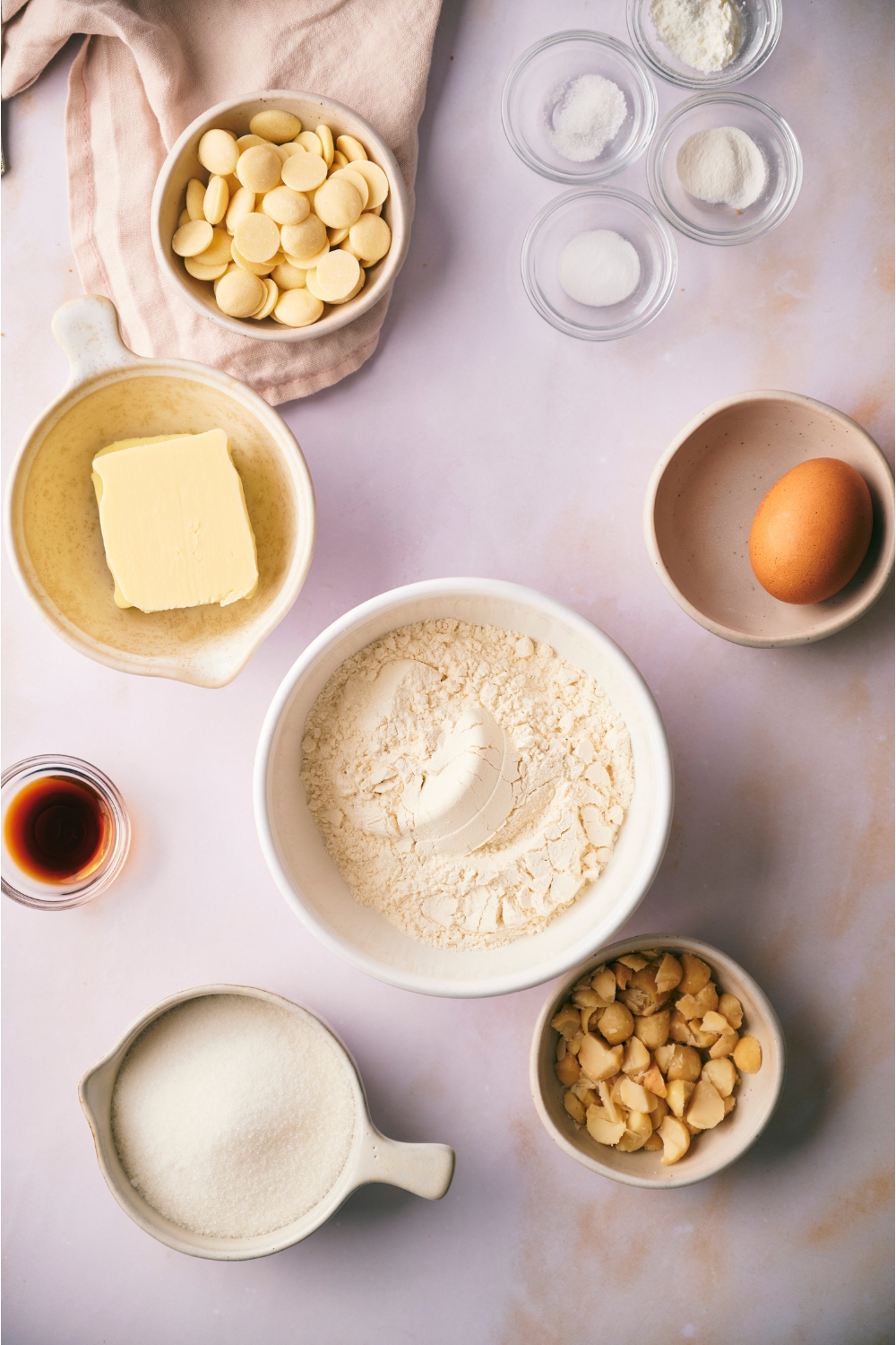 An assortment of ingredients including bowls of flour, white chocolate chips, an egg, vanilla extract, butter, and sugar.