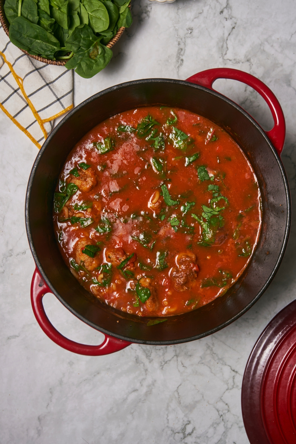 Red Dutch oven filled with meatballs in a tomato sauce and spinach has been added to the pot.