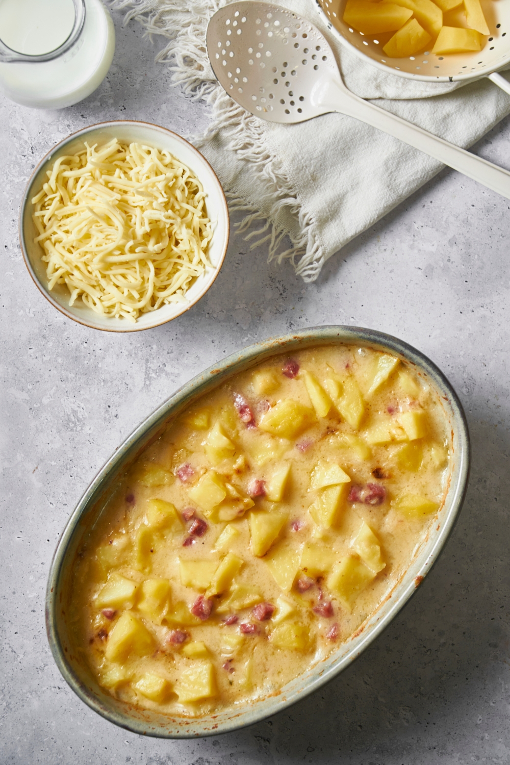 Ham and potato casserole in a large casserole dish, with a bowl of shredded cheese next to the casserole dish.