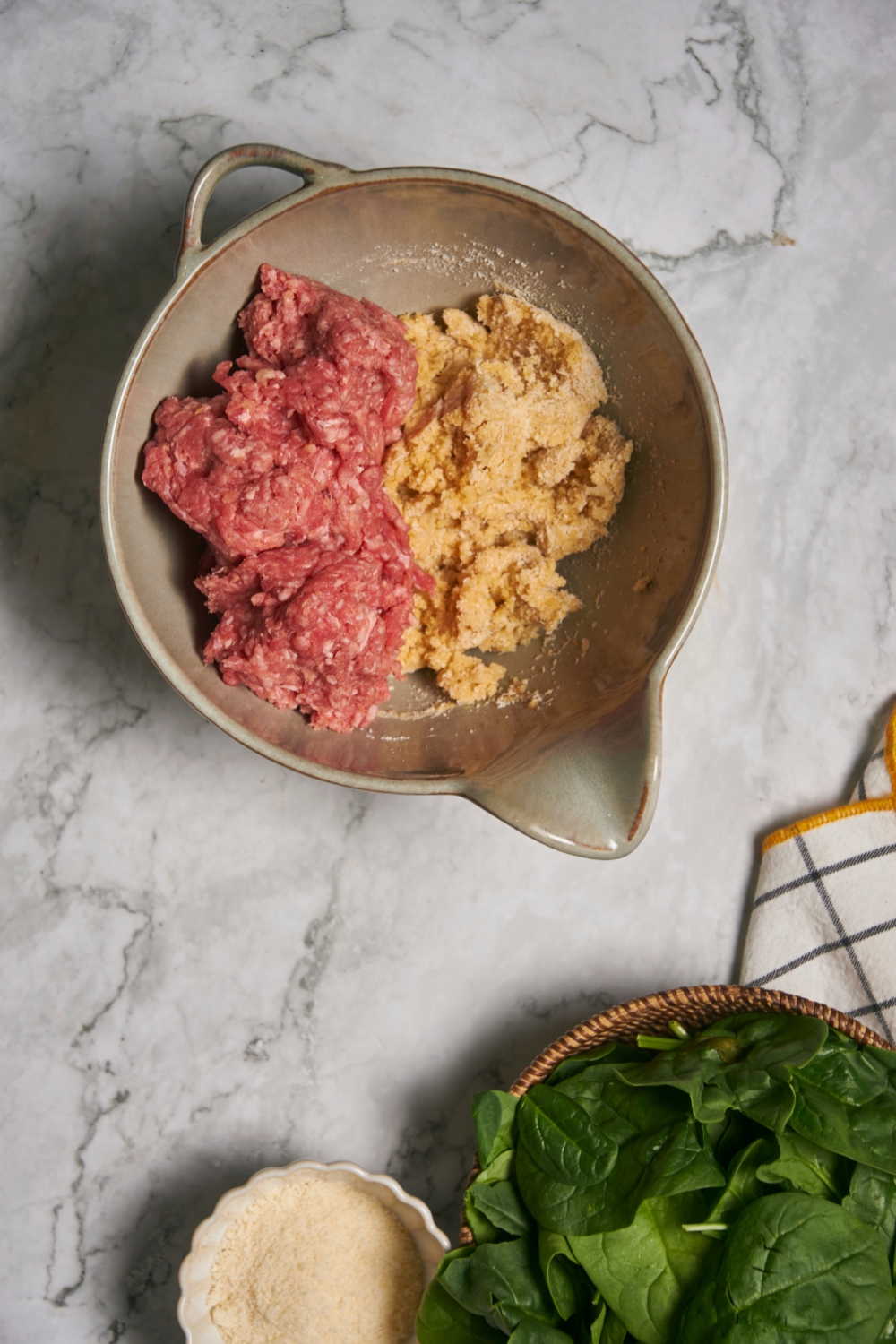 A mixing bowl filled half with raw meat and half with a bread crumb mixture, with a bowl of raw spinach and a small bowl of breadcrumbs next to the mixing bowl.
