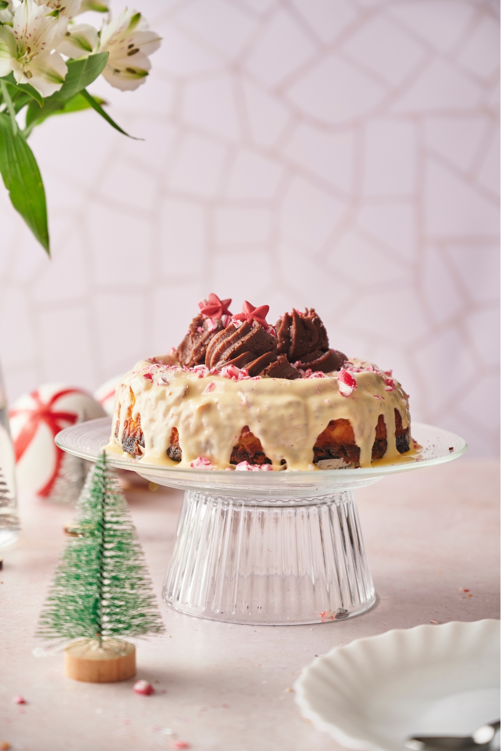 Peppermint cheesecake topped with white frosting, mounds of chocolate frosting, crushed candy canes, and star sprinkles. The cheesecake is on a clear cake stand.