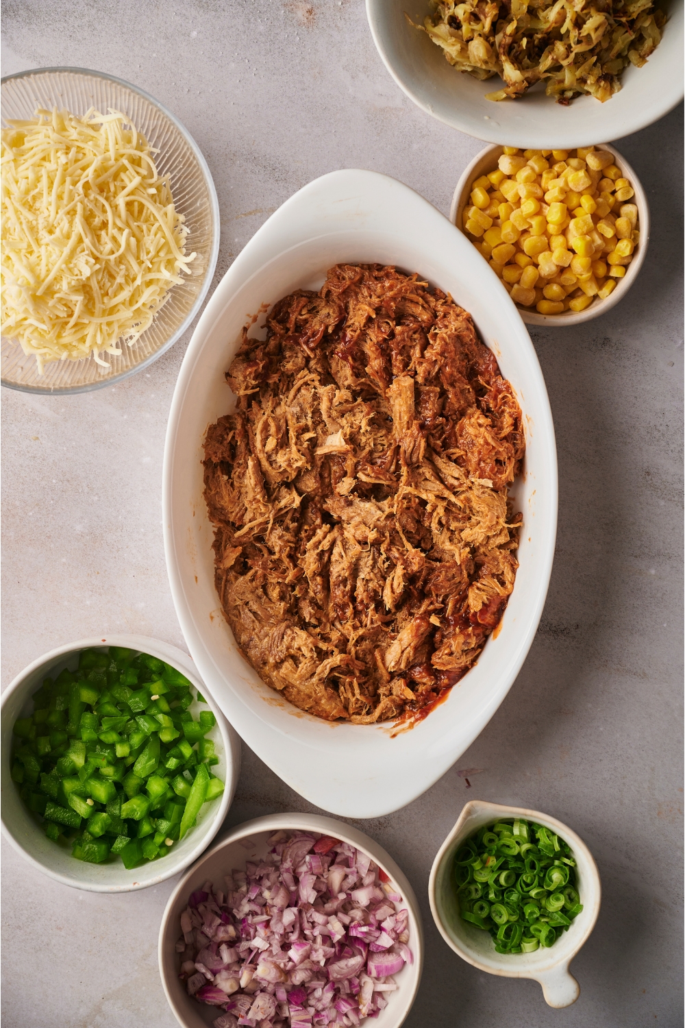 A white baking dish filled with BBQ pulled pork surrounded by an assortment of ingredients including bowls of corn, diced onion, diced peppers, shredded cheese, and diced green onion.