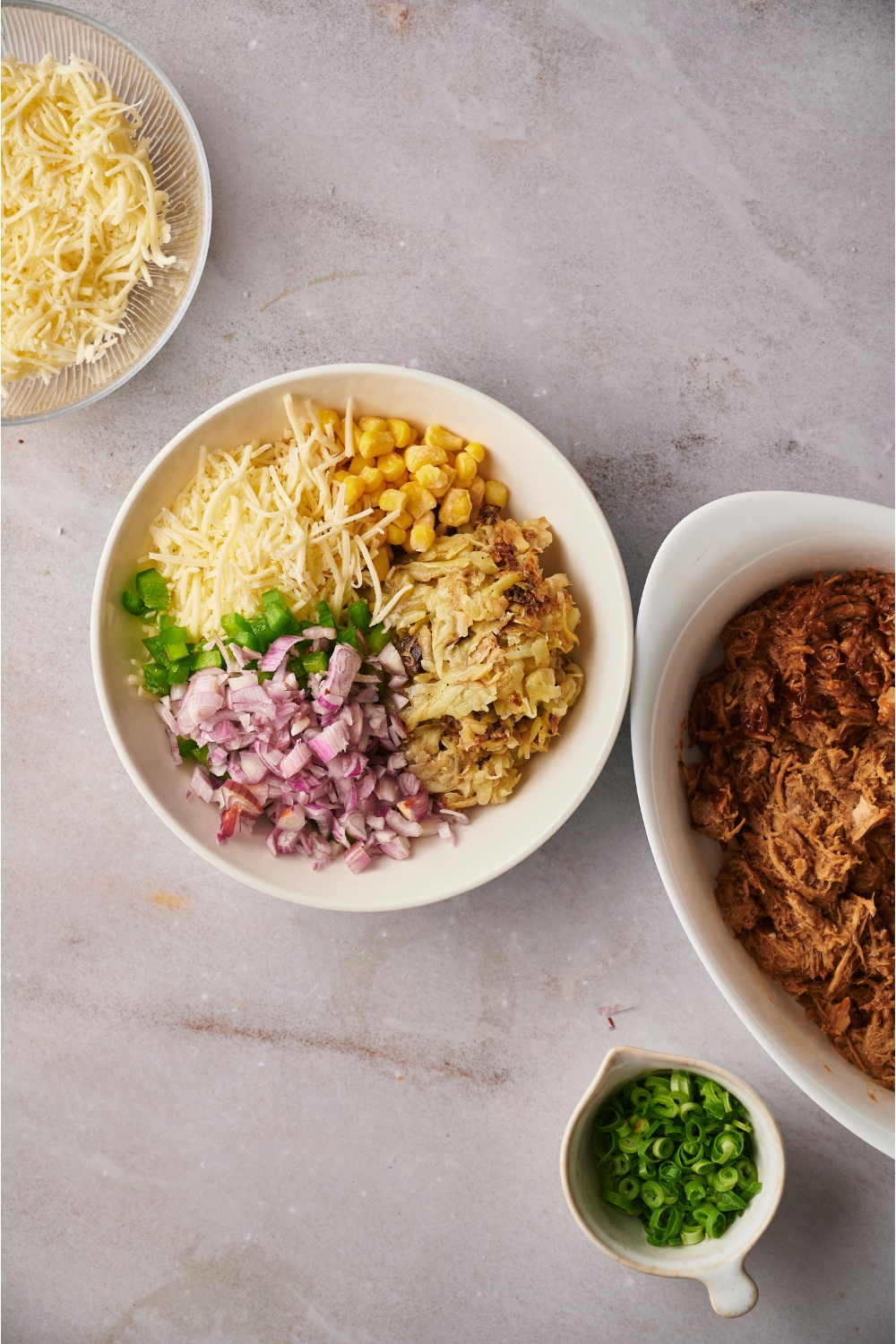White bowl filled with shredded hash browns, diced onion, diced bell peppers, corn, and shredded cheese. Next to the bowl is a white baking dish filled with pulled pork, a small dish of green onions, and a bowl of shredded cheese.