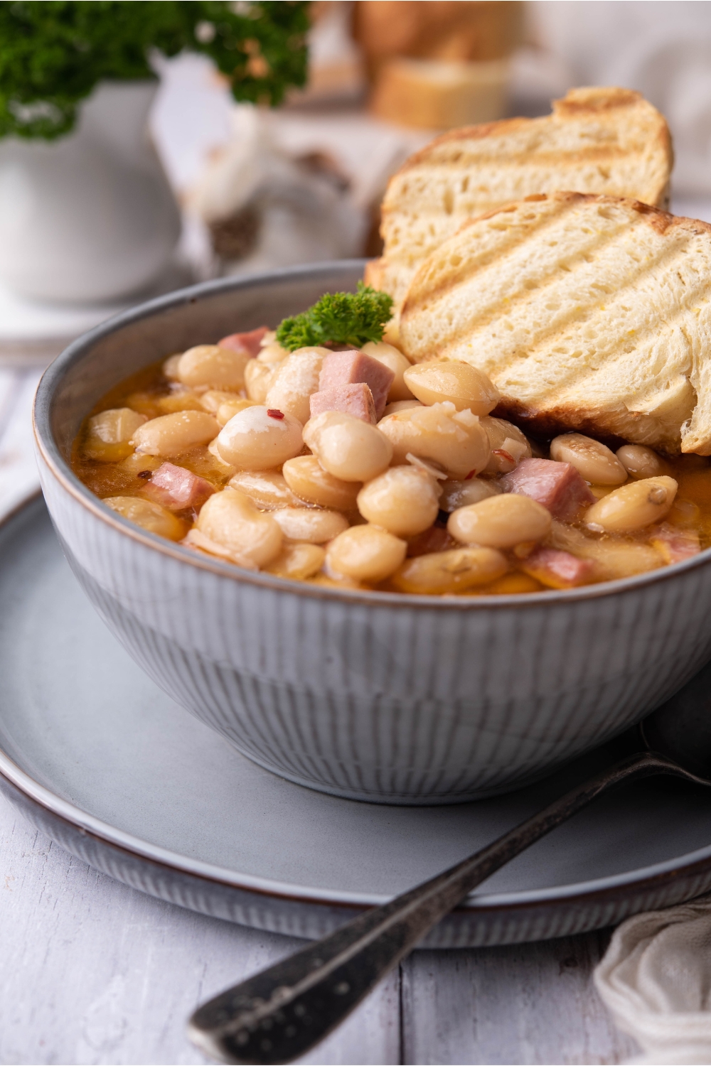 Butter beans with diced ham and two slices of toast on a blue bowl on top of a blue plate, with a spoon on the plate.