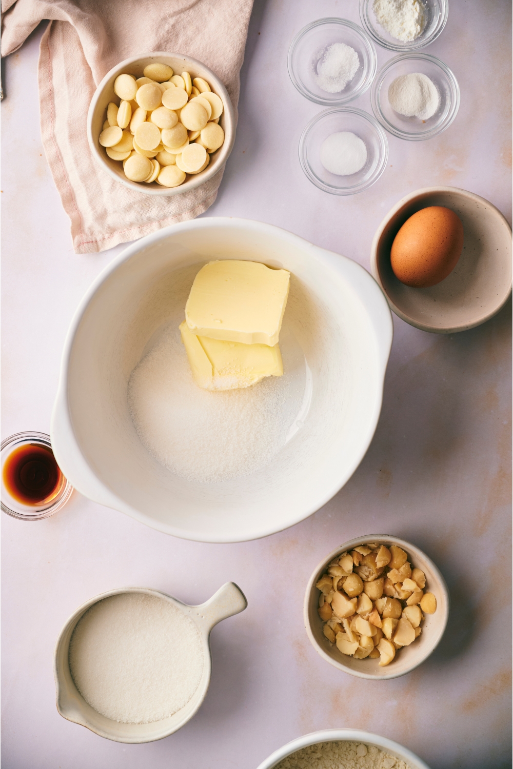 A white mixing bowl with butter and sugar in the bowl, not yet mixed together. The bowl is surrounded by ingredients including white chocolate chips, macadamia nuts, and vanilla extract.