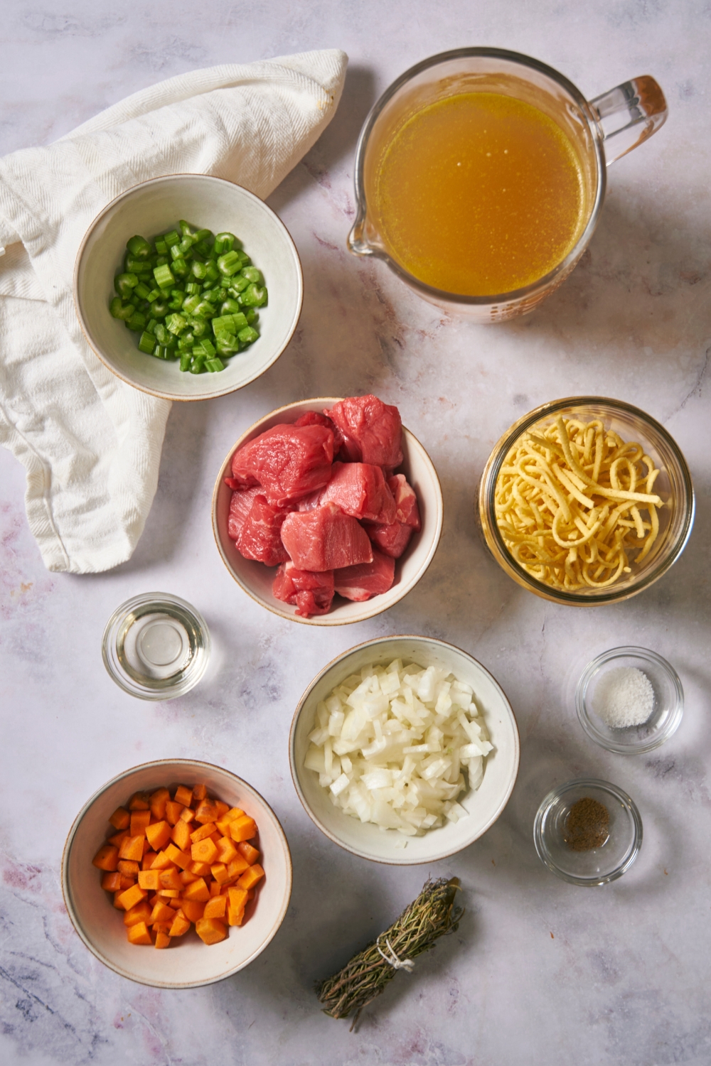 A bowl of broth, a bowl of diced green pepper, a bowl of beef cubes, a bowl of egg noodles, a bowl of onion, and a bowl of diced carrots.