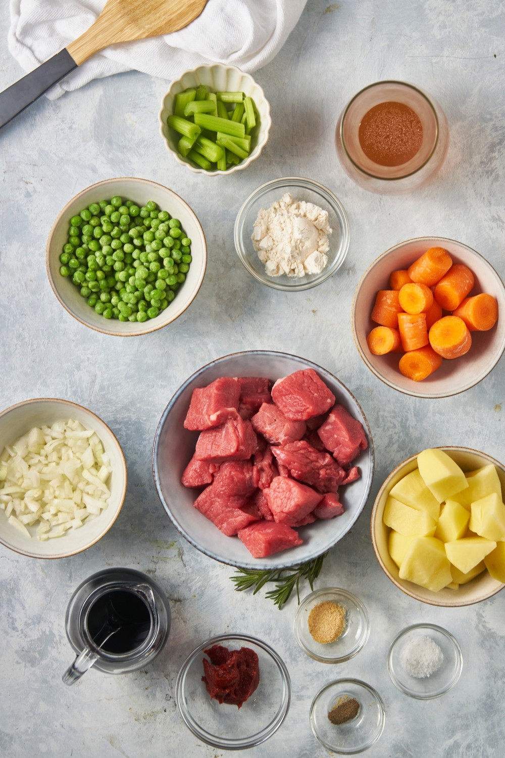A bowl of stew meat pieces, a bowl of chopped carrots, a bowl of potato chunks, and a bowl of peas all on a grey counter.