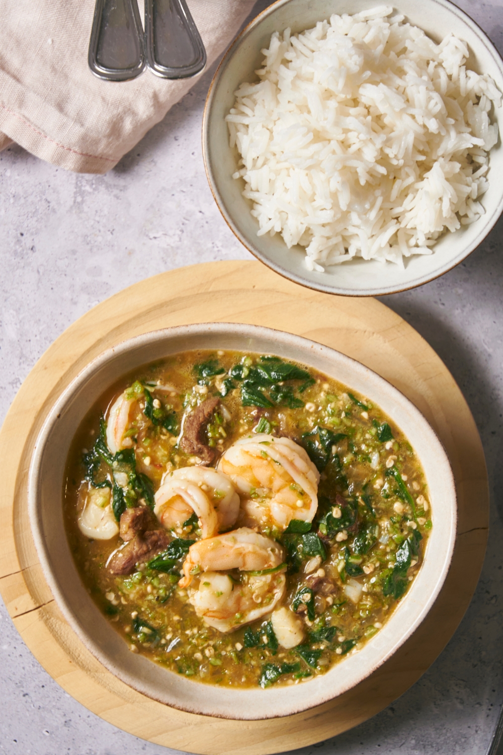 Shrimp on top of spinach and ground okra in a bowl filled with broth. Behind it is a bowl of white rice.