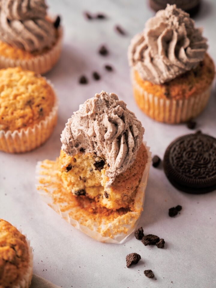 A Oreo cupcake that has a bite out of it on an unwrapped cupcake wrapper with piped frosting on top of the cupcake.