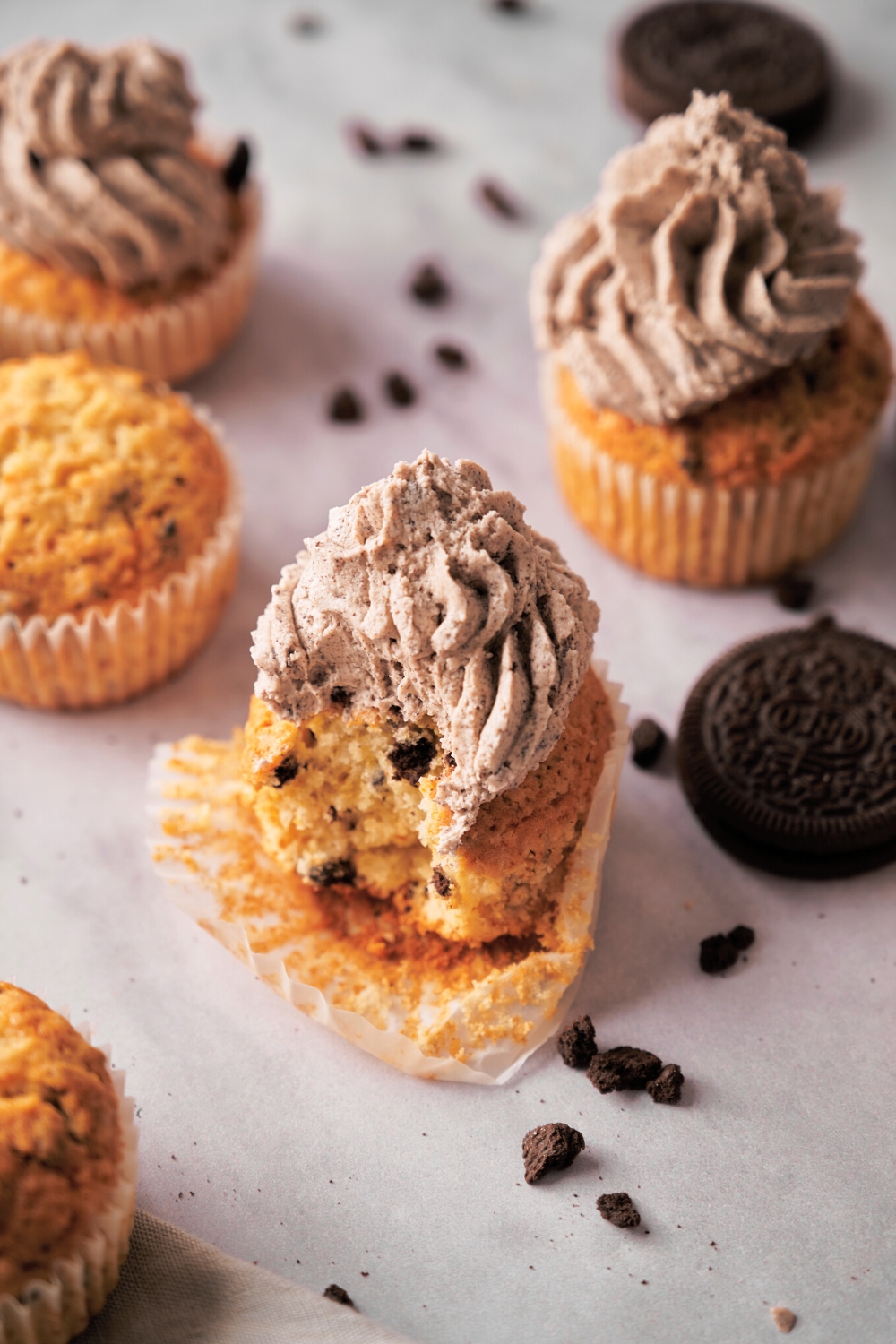 A Oreo cupcake that has a bite out of it on an unwrapped cupcake wrapper with piped frosting on top of the cupcake.