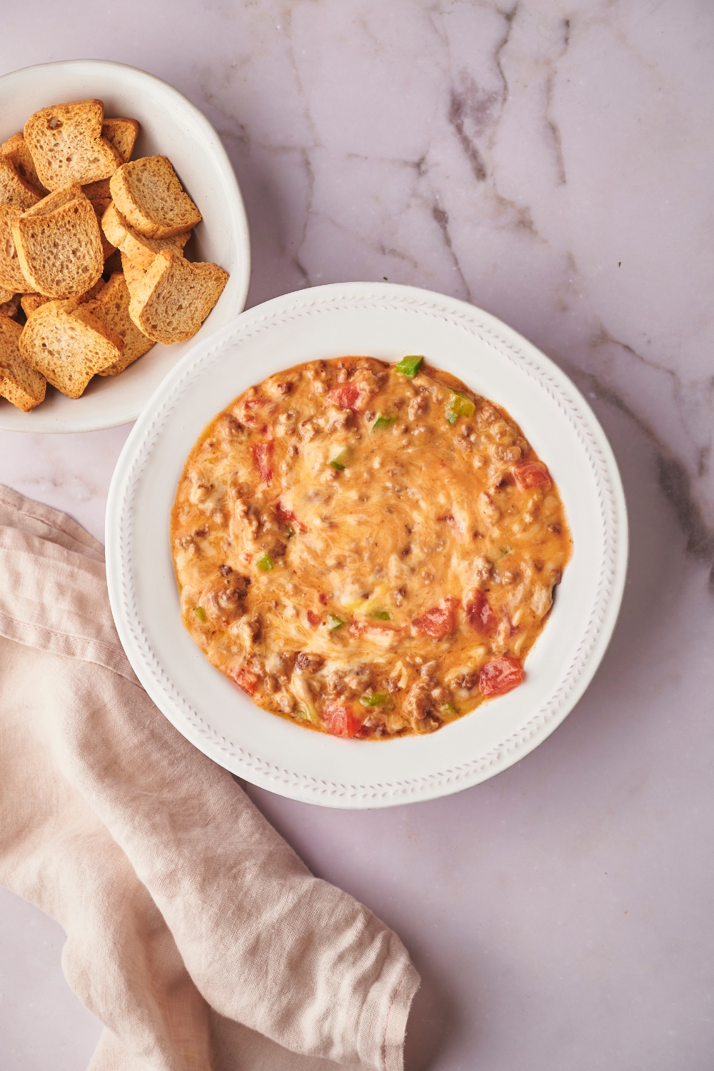 Rotel dip that is in a white bowl on a white counter. Behind the bowl is part of a bowl of mini toasts.