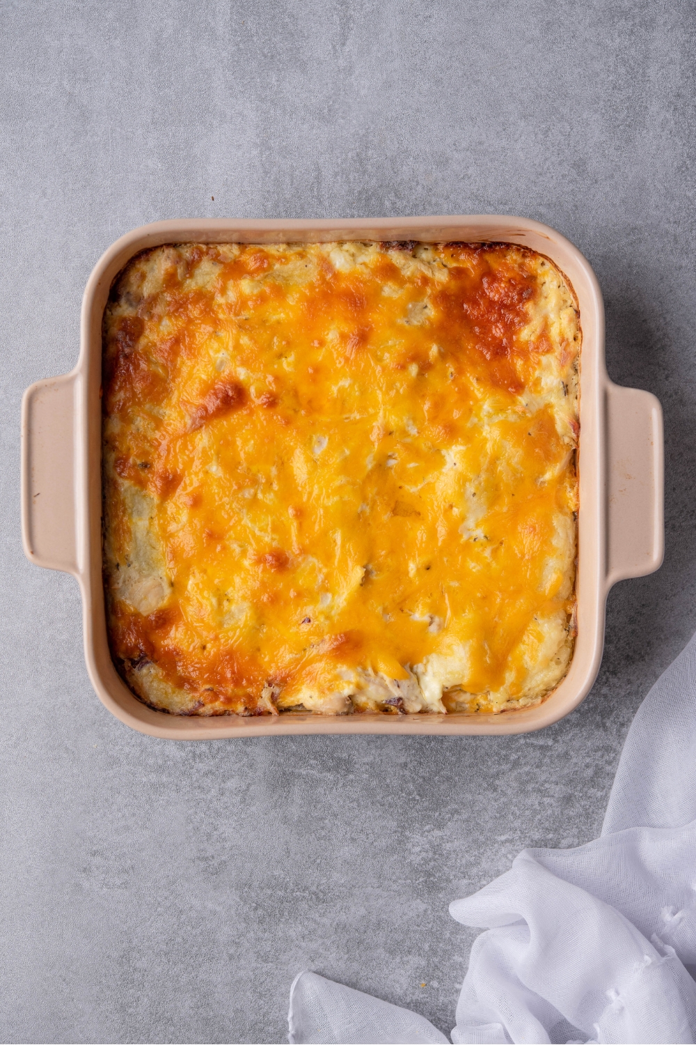 Melted shredded cheddar cheese on top of a casserole and a square casserole dish and a gray counter.