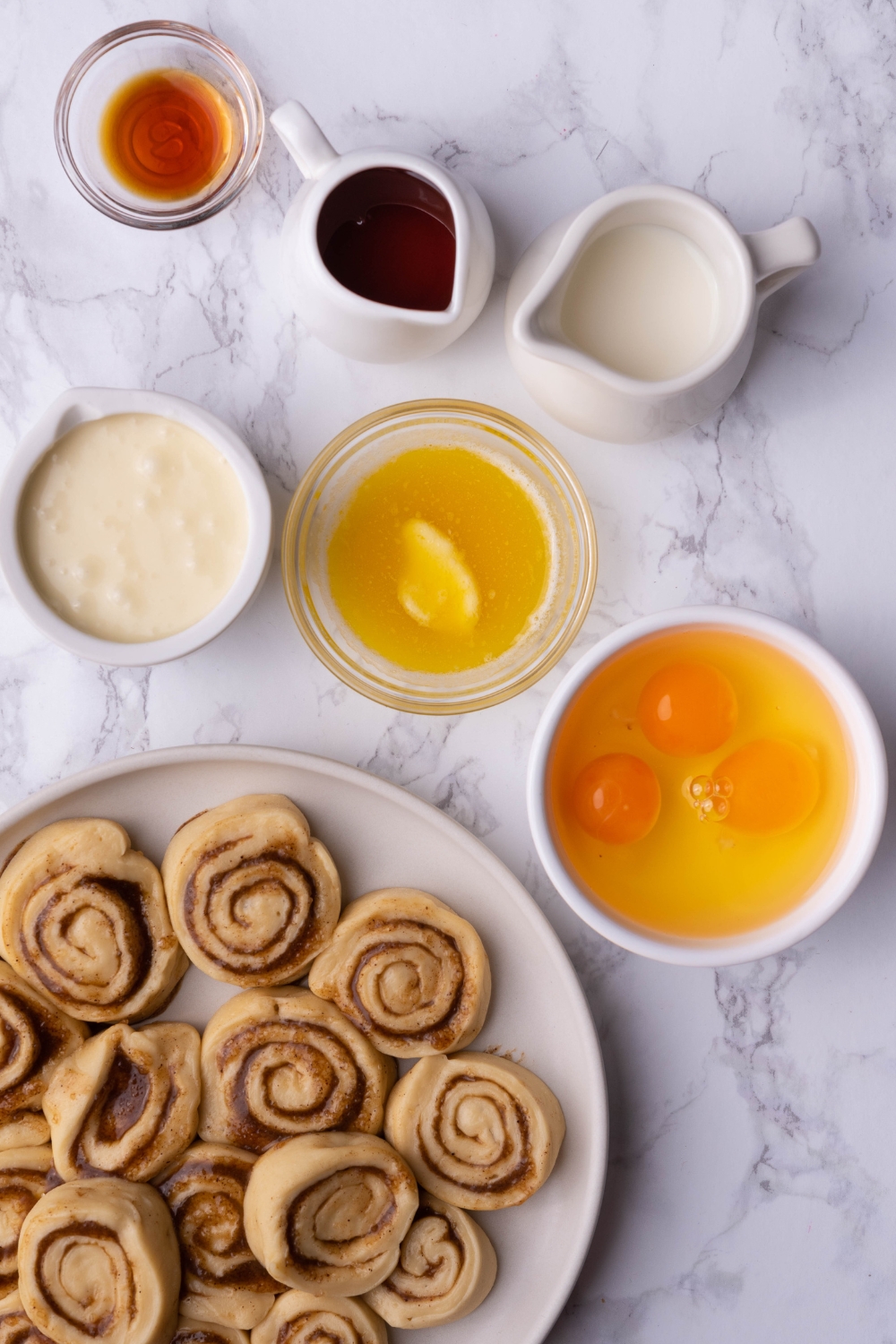 A bowl of maple syrup, a bowl of vanilla extract, a bowl of eggs, a bowl of malted butter, and part of a plat with cinnamon rolls on it.