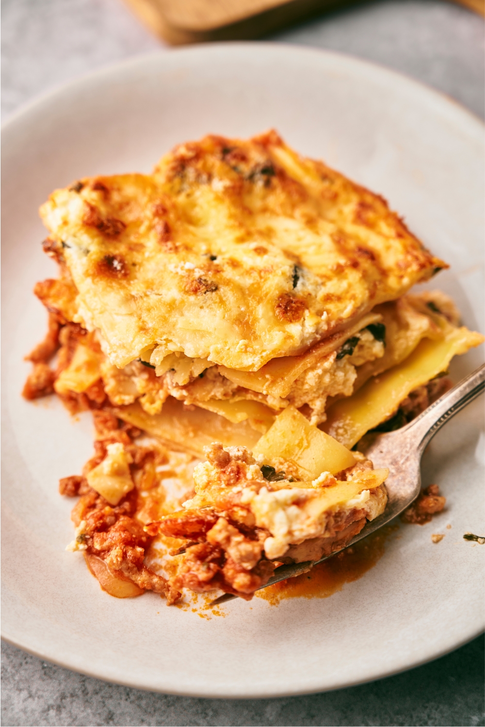 A square of beef lasagna on a white plate with a fork with some of the lasagna in front of it.